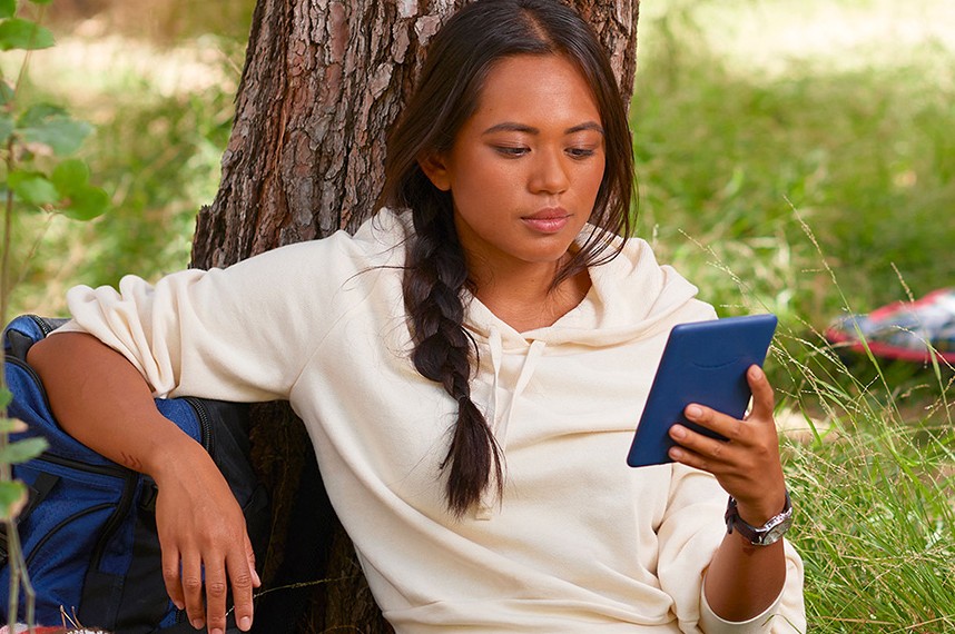 Pembaca bersandar di pohon sambil membaca Kindle biru